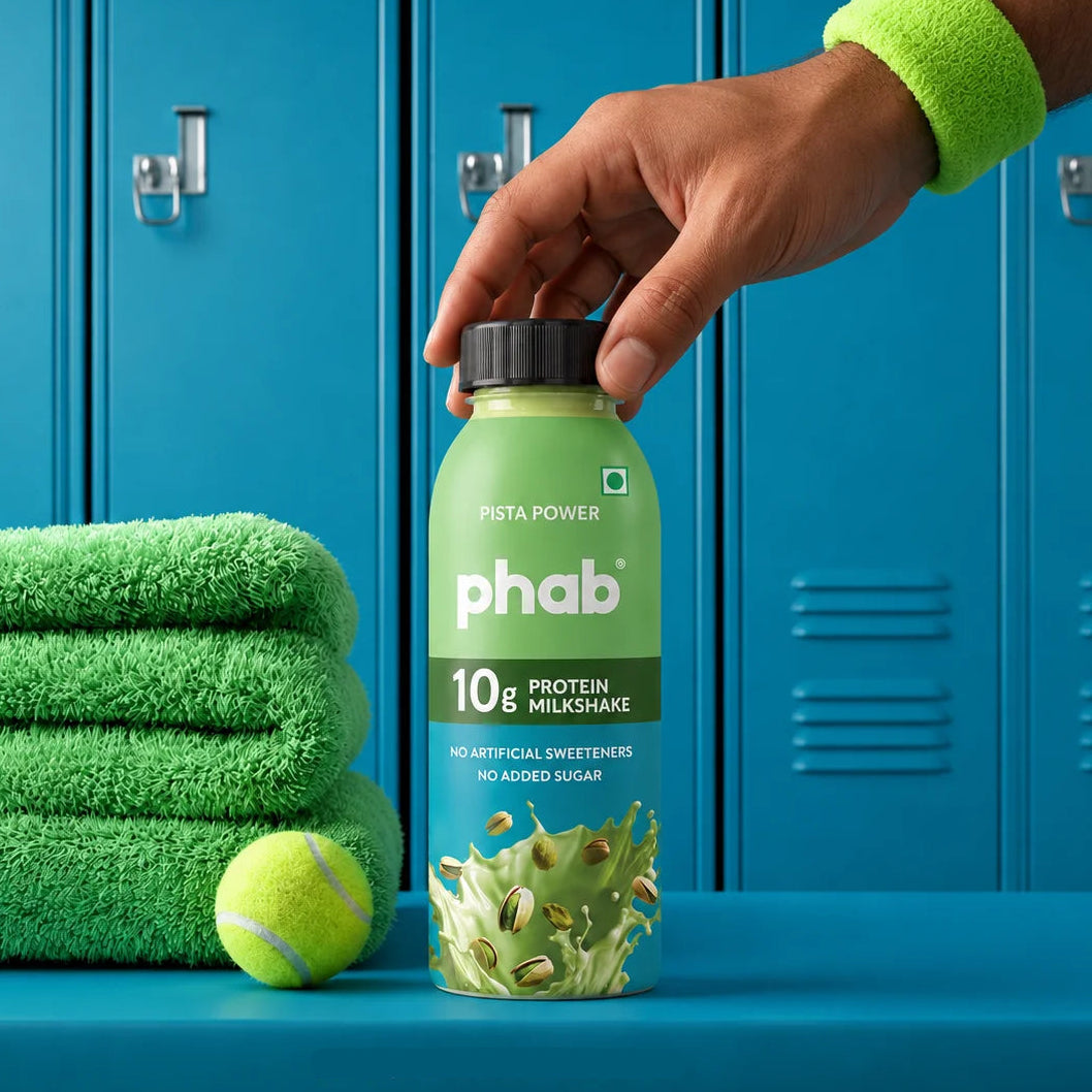 A hand wearing a pista green wristband holds a bottle of phab 10g Pista Power protein milkshake. The scene is set against a background of blue lockers, with a stack of green towels and a tennis ball on a blue surface in the foreground.